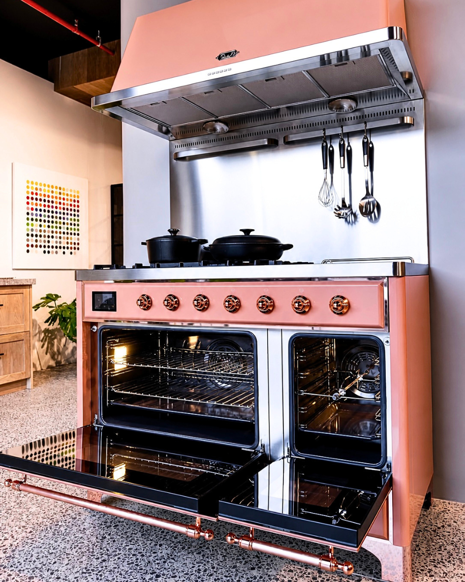 Custom pink ILVE range with matching hood, copper trim, open double ovens, and cookware in a modern kitchen.